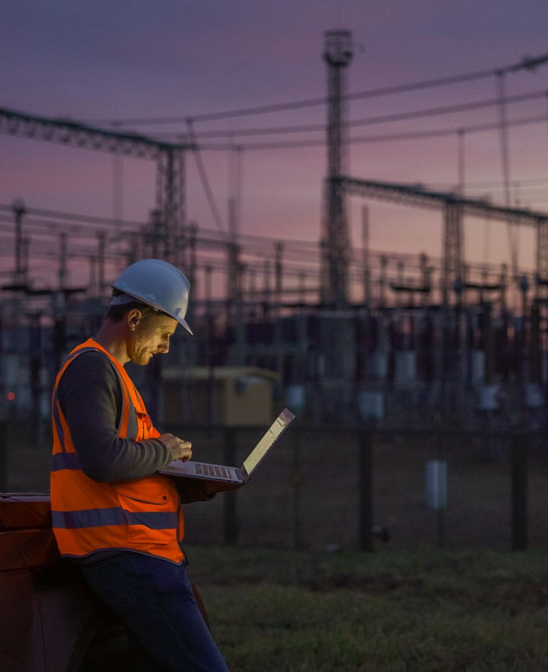 Ein Ingenieur der Energieversorgung in Schutzhelm und Warnweste beobachtet in der Abenddämmerung die Anzeigen auf einem Laptop in einem Umspannwerk.