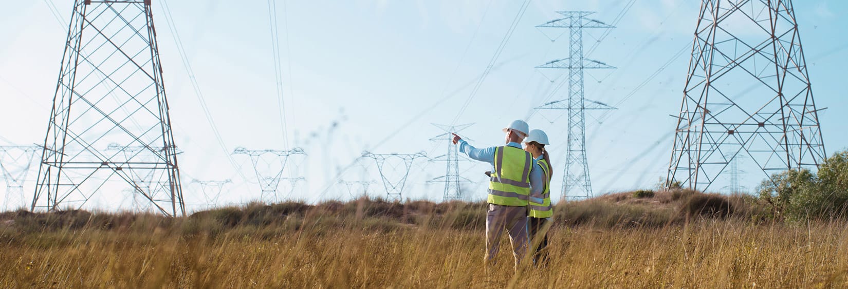 Zwei Arbeiter eines Energieversorgers in Sicherheitswesten und mit Helmen inspizieren Strommasten auf einer Wiese.